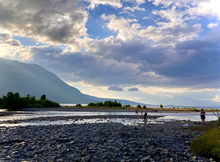 Bild: en stenig strand med berg i fonden, några personer vadar i vattnet.