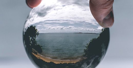 En delvis molning strandvy ses genom en glaskula som hålls mellan tummen och pekfingret i någons hand. 
