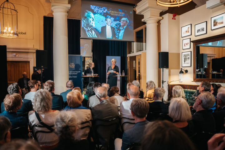 Bild: perspektiv över publikhav mot scen med Anna och Sofia, backdrop bild med porträtt av Torsten i överkant